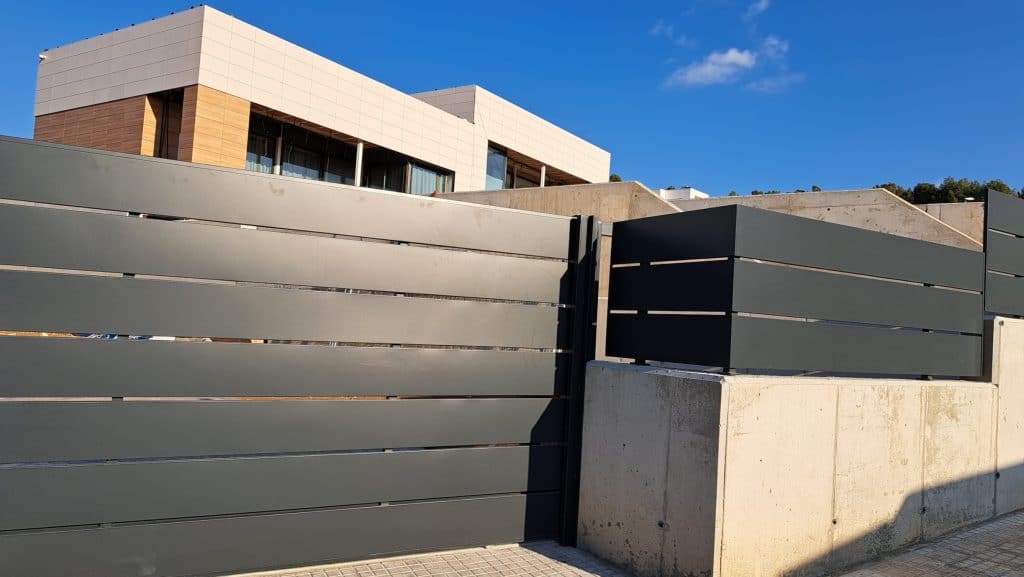 Casa moderna con amplios ventanales, situada tras una alta valla gris de hormigón y soluciones metálicas, captada en un día soleado.
