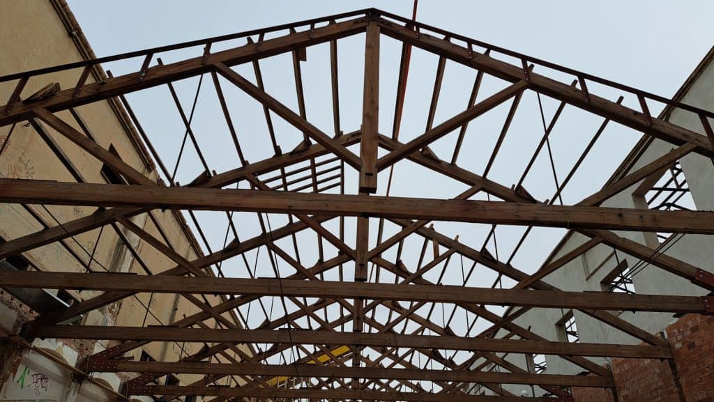 Vigas de madera del tejado de un edificio en construcción o en proceso de renovación, con el cielo a la vista a través de la estructura y paredes parciales a ambos lados; ideal para integrar soluciones metálicas que aporten mayor resistencia y durabilidad.