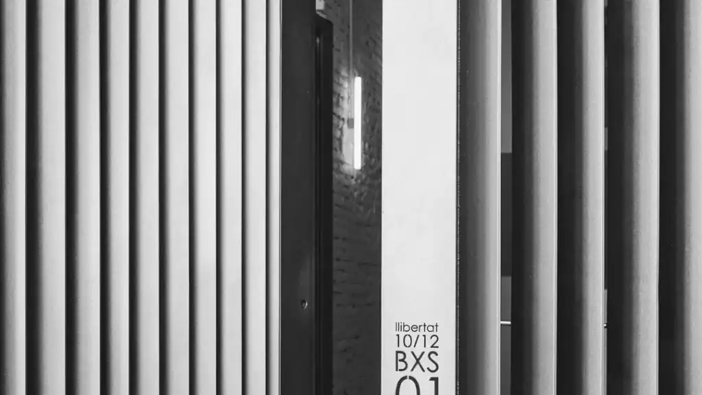 Fotografía en blanco y negro de unas barras metálicas dispuestas verticalmente, con un estrecho hueco que deja entrever un pasillo iluminado; en una de las barras se lee el texto «llibertat 10/12 BXS 01». Parece la entrada de una cerrajería metálica