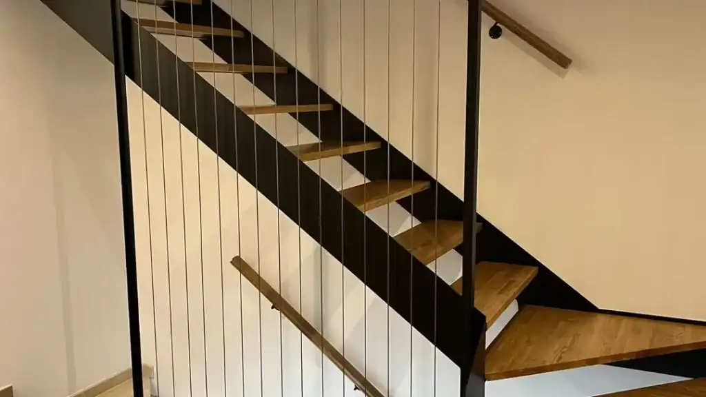Escalera moderna con peldaños de madera, estructura de metal negro y cables finos verticales a modo de barandilla, que destaca por sus elegantes soluciones metálicas sobre una pared blanca.