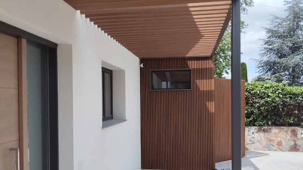Una moderna entrada exterior que cuenta con una pared blanca, una ventana con marco negro, una pared de lamas de madera y un techo de pérgola de madera horizontal realzado con soluciones metálicas para mayor durabilidad y estilo.