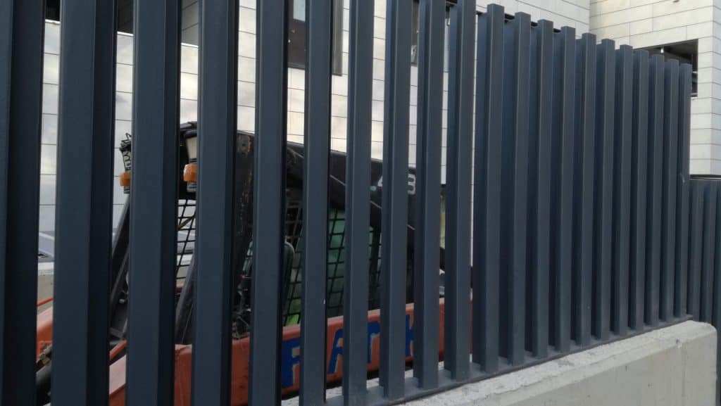 Una valla metálica con barras verticales —que forma parte de «soluciones metálicas innovadoras»— oculta parcialmente un vehículo de obra aparcado detrás de ella, cerca de un edificio moderno.